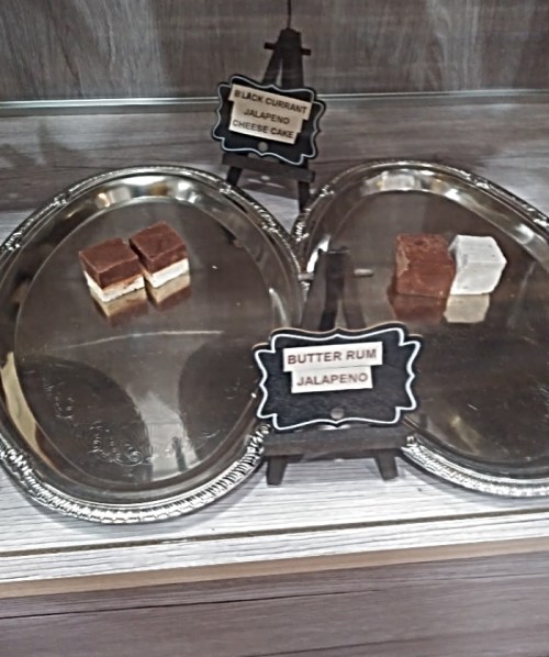 Two silver trays display small dessert squares labeled "Black Currant Jalapeño Cheesecake" and "Butter Rum Jalapeño."