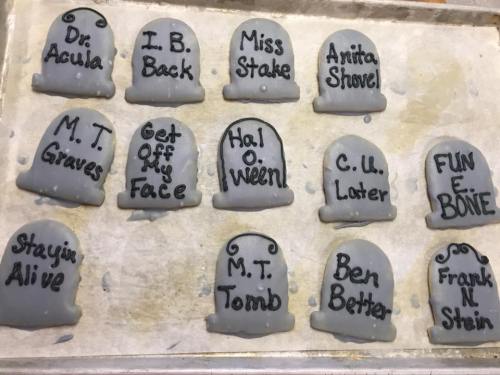 A tray of Halloween-themed cookies shaped like tombstones with humorous names written on them.