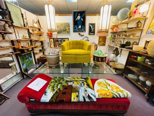 A vintage shop interior featuring a yellow chair, a red bench with a blanket, and various decorative items on shelves.