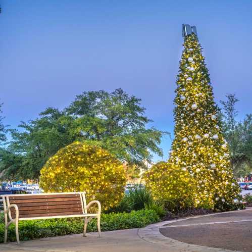 A festive tree adorned with lights stands in a park, with a bench nearby and greenery in the background.