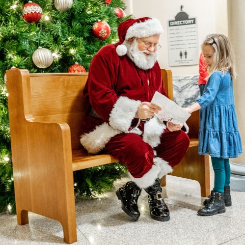 Santa Claus sits on a bench, reading a letter from a young girl in a blue dress, with a decorated Christmas tree nearby.
