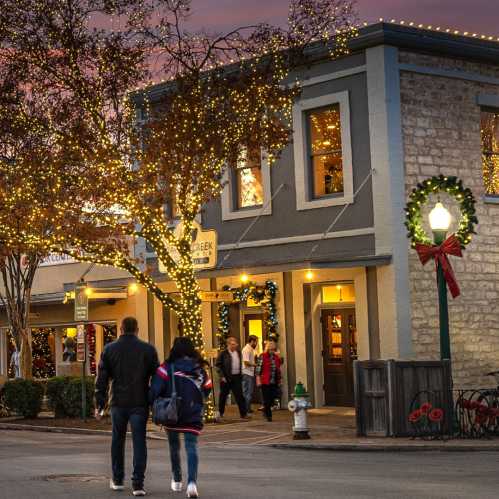 Couple walking past a festive building adorned with holiday lights and wreaths, under a colorful sunset sky.