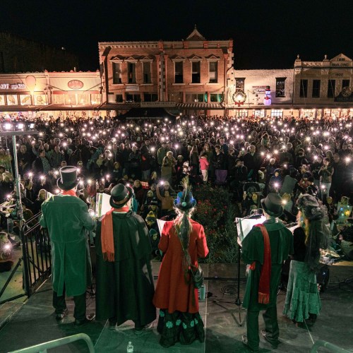 A large crowd in costumes gathers at night, holding lights, with a stage and historic buildings in the background.