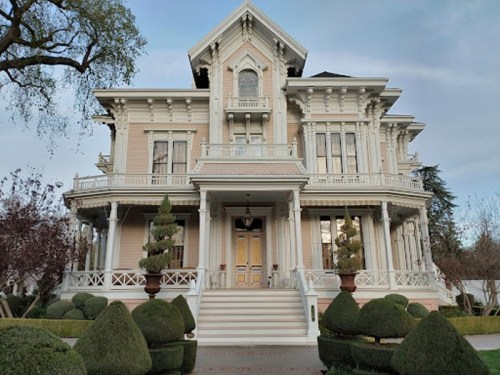 A large, ornate Victorian-style house with a pink exterior, intricate details, and manicured hedges in front.