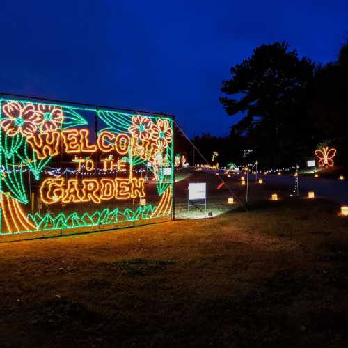 A brightly lit sign reading "Welcome to the Garden" surrounded by decorative lights and flowers at dusk.