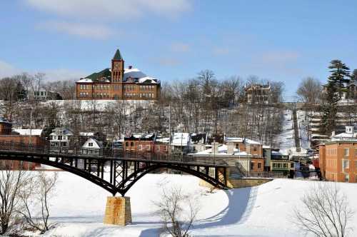 A snowy landscape featuring a historic building on a hill, with a bridge in the foreground and trees in the background.