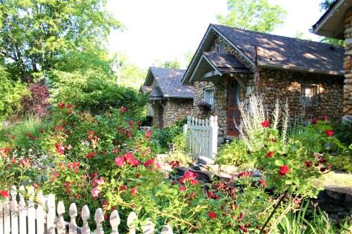 A charming stone cottage surrounded by vibrant flowers and greenery, with a white picket fence in the foreground.