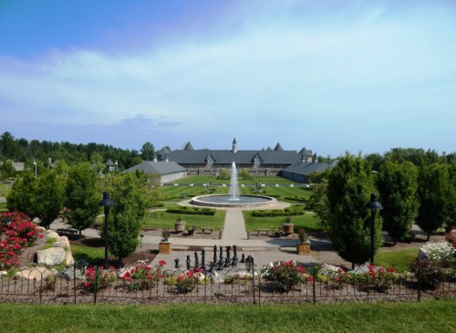 A landscaped garden with a fountain, surrounded by trees and flowers, leading to a large building under a blue sky.
