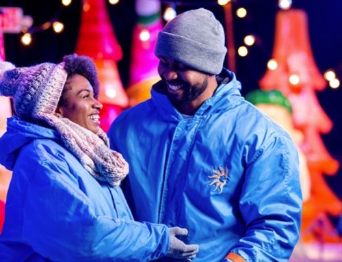 A couple in winter jackets smiles at each other, surrounded by colorful lights and festive decorations.