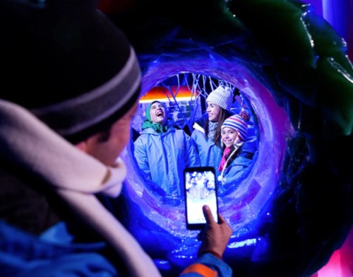 A person takes a photo of three smiling kids in winter attire, framed by colorful ice sculptures.