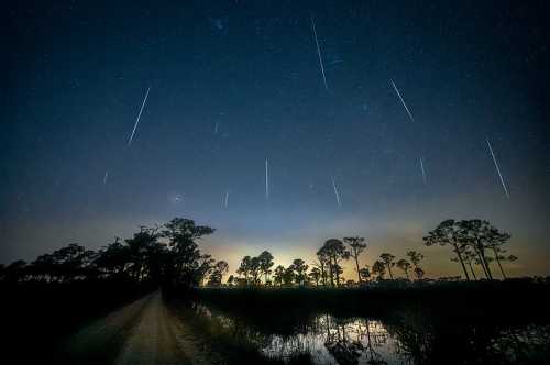 A starry night sky with multiple meteor trails above a dark landscape and a reflective water body.