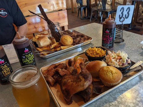 A tray of barbecue featuring chicken, ribs, corn, coleslaw, and muffins, with drinks and a table number in the background.