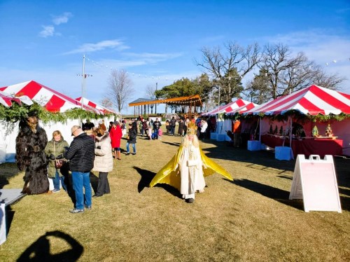 A festive outdoor market with red and white tents, people browsing, and a person in a yellow costume walking by.