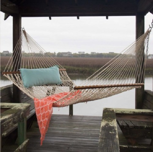 A cozy hammock with a blue pillow and coral throw, set on a wooden dock overlooking a calm marsh.