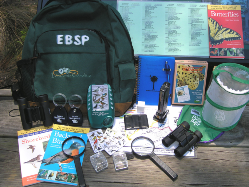 A green backpack with binoculars, field guides, a magnifying glass, and other nature exploration tools on a table.