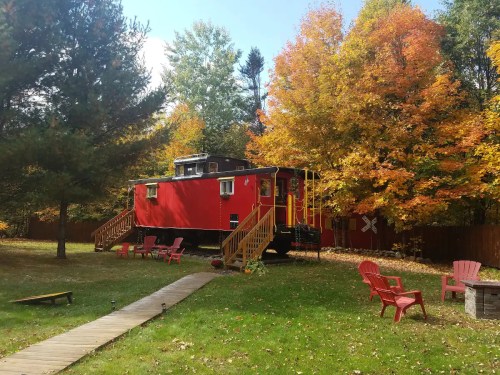 A red train caboose in a colorful autumn setting, surrounded by trees and a grassy area with chairs.