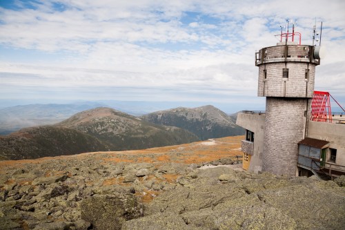 A stone tower with antennas overlooks rocky terrain and distant mountains under a cloudy sky.