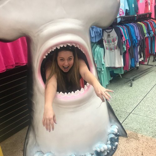 A woman playfully poses inside a large shark mouth display, surrounded by colorful clothing racks.