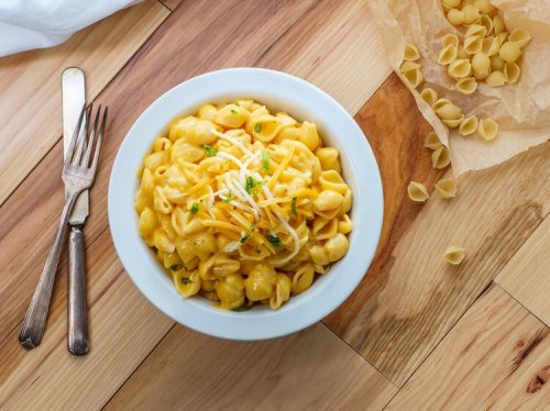 A bowl of creamy macaroni and cheese topped with shredded cheese, surrounded by uncooked pasta on a wooden table.