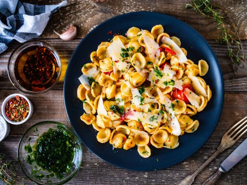 A plate of pasta with herbs, cheese, and red peppers, surrounded by garlic, spices, and olive oil on a wooden table.