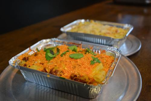 Two aluminum trays of baked dishes, one topped with orange crumbs and cilantro, the other creamy and cheesy.