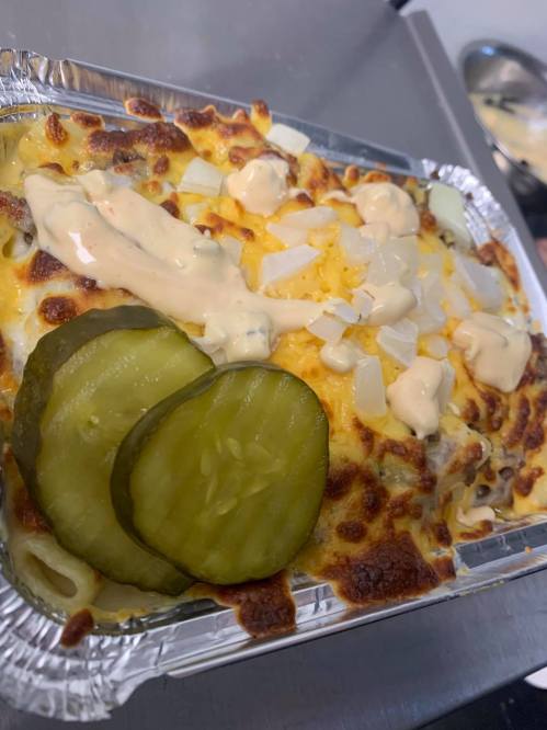 A baked dish topped with melted cheese, pickles, and a creamy sauce, served in a foil container.