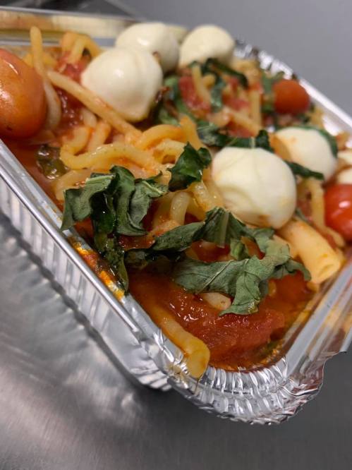 A close-up of a pasta dish with tomatoes, mozzarella balls, and greens in a clear takeout container.