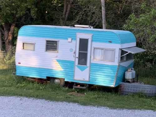 A vintage turquoise and white camper trailer parked on grass, surrounded by trees and gravel.