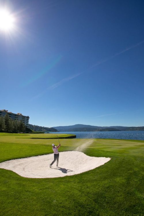 A golfer swings from a sand trap on a sunny day, with a lake and mountains in the background.