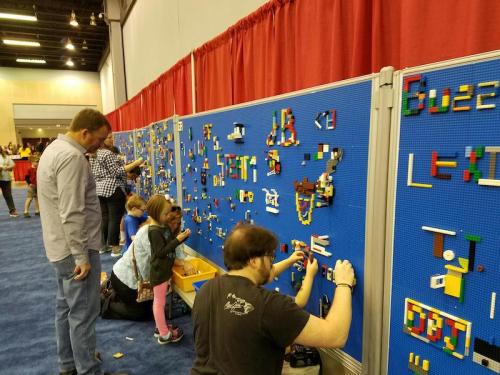 A group of people, including children, building with colorful LEGO bricks on a blue wall at an event.