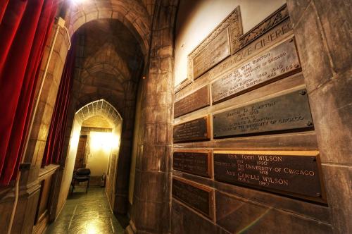 A dimly lit hallway with stone walls, red curtains, and plaques commemorating individuals on the right.