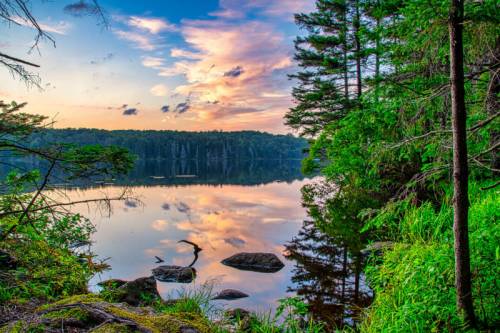 Serene lake at sunset, surrounded by trees and rocks, with colorful clouds reflecting on the water's surface.