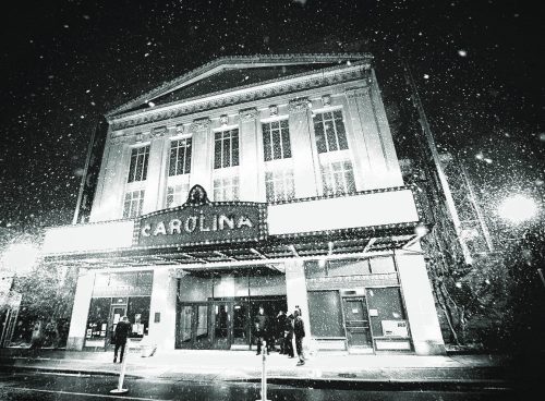 A historic theater, "Carolina," illuminated at night with snow falling and people gathered outside.