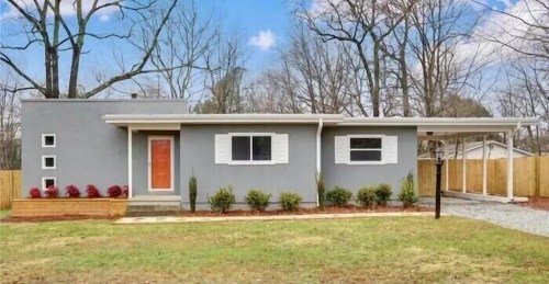 A modern gray house with a red door, white shutters, and a carport, surrounded by a grassy yard and trees.