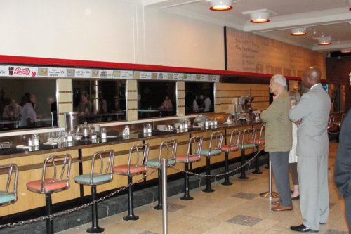 A vintage diner counter with empty stools and a mirrored wall, featuring patrons observing the space.