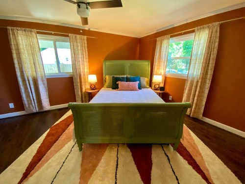 Cozy bedroom with a green bed, warm orange walls, two windows, and a patterned rug. Soft lighting from bedside lamps.