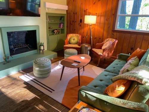 Cozy living room with a green sofa, orange chairs, a coffee table, and a fireplace, surrounded by wooden walls and natural light.