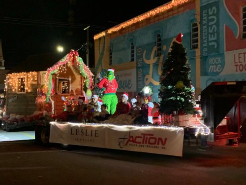 A festive parade float featuring the Grinch, Christmas trees, and children in Santa hats, decorated with lights.