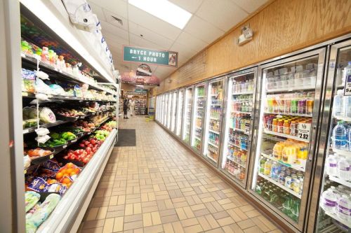 A grocery store aisle with fresh produce on one side and refrigerated drinks on the other, under bright lighting.