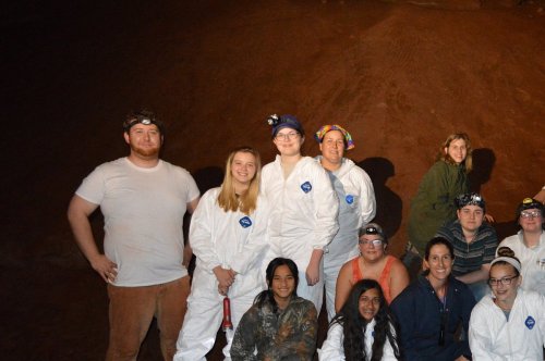 A group of people in white jumpsuits and headlamps poses together in a cave-like setting.