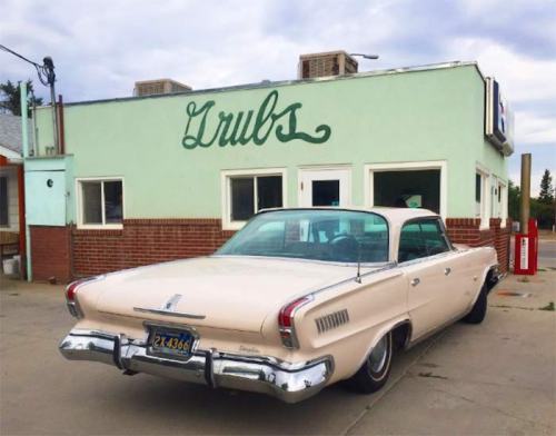 A vintage pink car parked in front of a retro diner with the sign "Grubs" on a green wall.