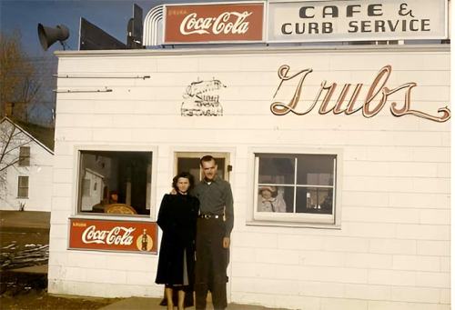 A man and woman stand outside a vintage café with a Coca-Cola sign, featuring a curb service window.