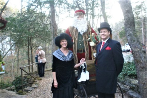 A man in a top hat and a woman in a black dress pose with a large Santa figure in a wooded area.