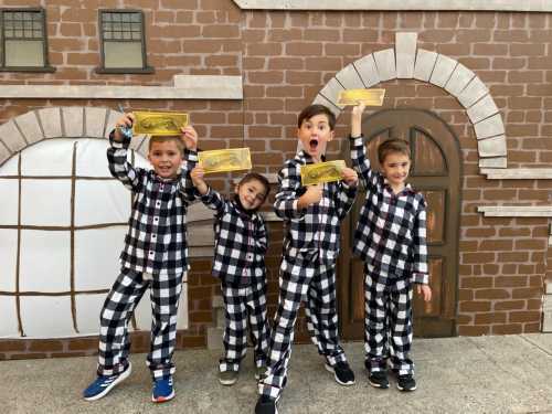 Four children in matching black and white checkered pajamas hold up golden tickets, smiling in front of a brick wall backdrop.