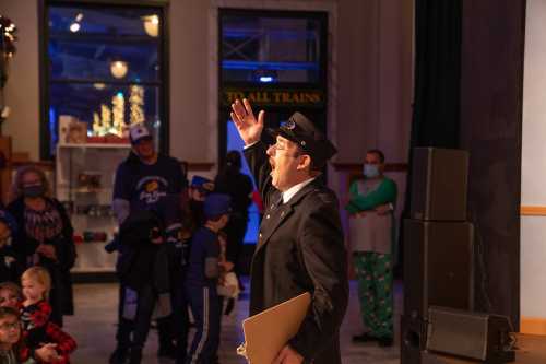 A performer in a conductor's outfit gestures while addressing an audience of children and families in a festive setting.