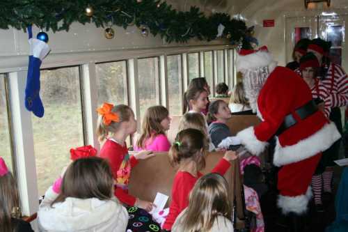 A festive train interior with children in holiday attire, interacting with Santa and an elf. Decorations adorn the windows.