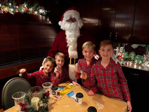 Four boys in matching plaid pajamas smile with Santa at a festive table decorated for Christmas.