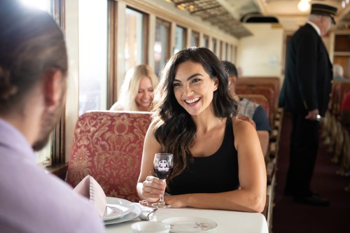 A woman smiles while holding a glass of wine at a dining table in a train car, with other diners in the background.