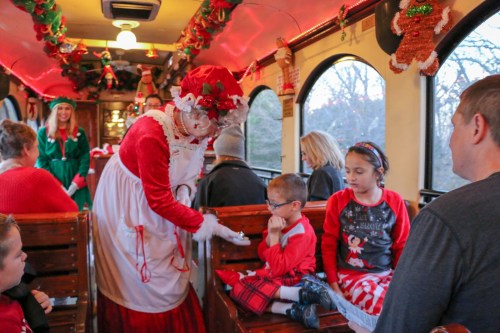 A festive train interior with children in holiday attire, as a character in a red dress interacts with them.
