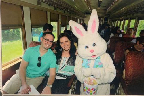 A couple smiles for the camera with a person in a bunny costume holding a basket on a train. Passengers are seated behind them.
